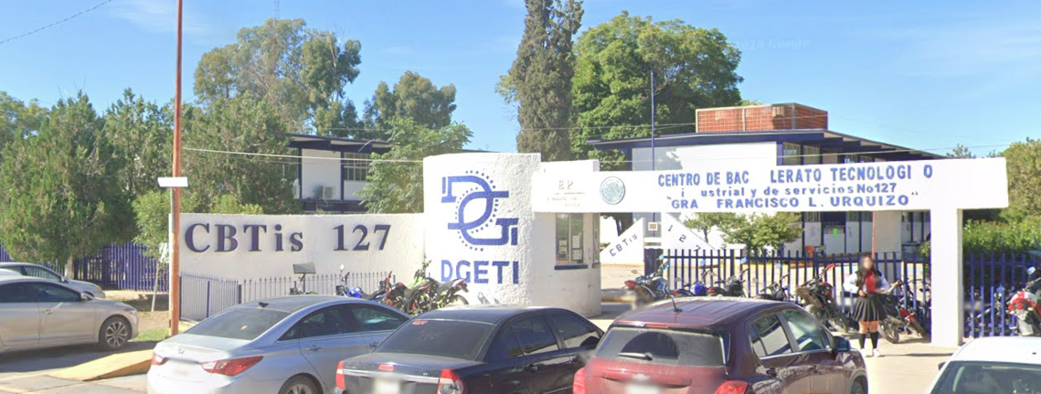 Fotografía en la que se muestra la entrada del plantel, conformada por muros de color blanco que en letras grandes azules dicen Cebetis 127. A la derecha de los muros hay una reja de color azul que da acceso al plantel.